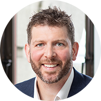 A middle-aged man with short, styled hair and a beard smiles at the camera. He wears a suit jacket over a white shirt. Large windows with natural light are visible in the background.