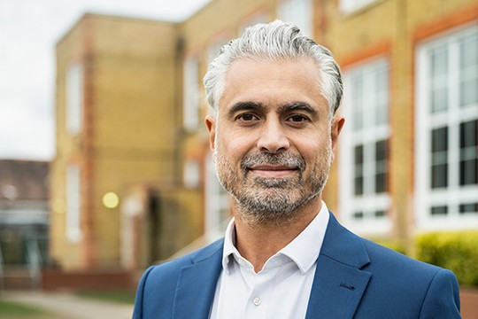 A man with gray hair and a beard wears a blue suit and white shirt. He stands confidently in front of a brick building with large windows and green shrubbery.