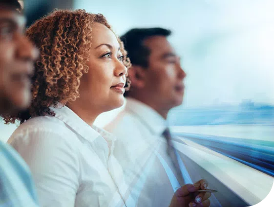 A woman with curly hair sits thoughtfully alongside two men, gazing forward as blurred motion suggests high-speed movement, conveying a sense of anticipation and focus in a dynamic environment.