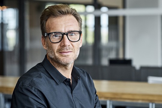 A man with short hair and glasses sits on a simple chair, looking directly at the camera. He is dressed in a black shirt, with a modern office setting in the background.