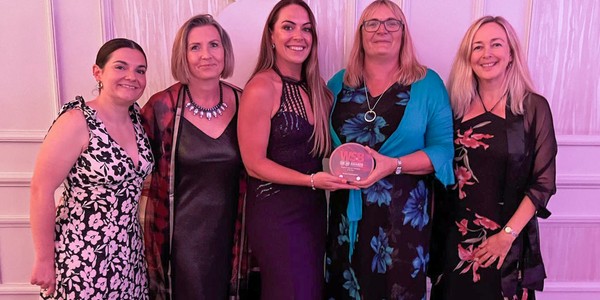 Five women pose together at a celebration event, with one holding an award. They wear elegant dresses, surrounded by a decorated backdrop. The atmosphere appears festive and lively.