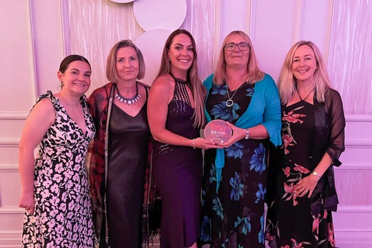 Five women pose together at a celebration event, with one holding an award. They wear elegant dresses, surrounded by a decorated backdrop. The atmosphere appears festive and lively.