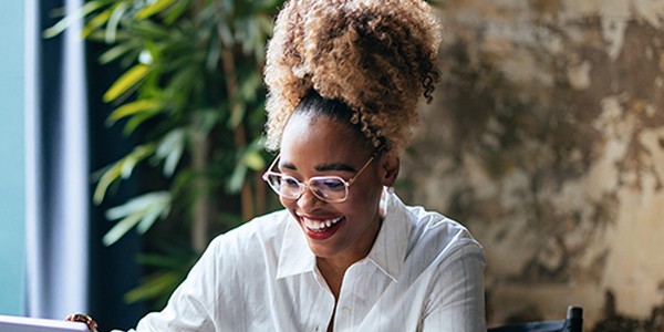 A woman with curly hair is smiling while using a laptop. Lush green plants surround her in a softly lit, textured indoor space, creating a relaxed atmosphere.