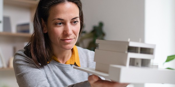 A woman holds a white architectural model of a building, examining it thoughtfully. She sits at a desk surrounded by indoor plants and a bright, modern workspace.