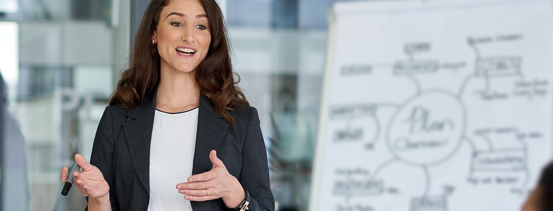 A woman in a suit gestures actively while presenting ideas on a whiteboard. The background features a modern office environment, indicating a professional meeting setting.
