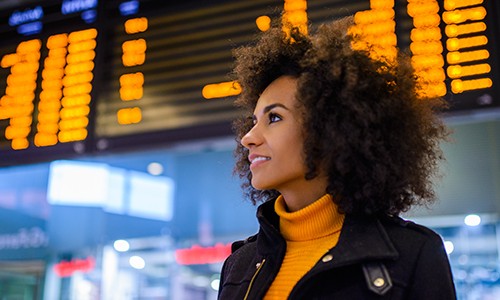 A young woman with curly hair is gazing upward, smiling, in an airport terminal. An electronic display board with flight information is illuminated behind her, creating an energetic travel atmosphere.