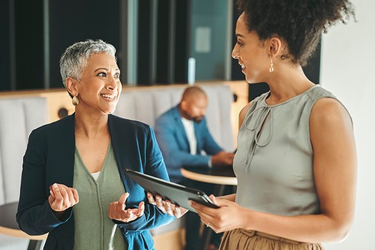 Two women converse happily, with one holding a tablet. In the background, a man focuses on a device. The setting appears modern and casual, suggesting a collaborative workspace.