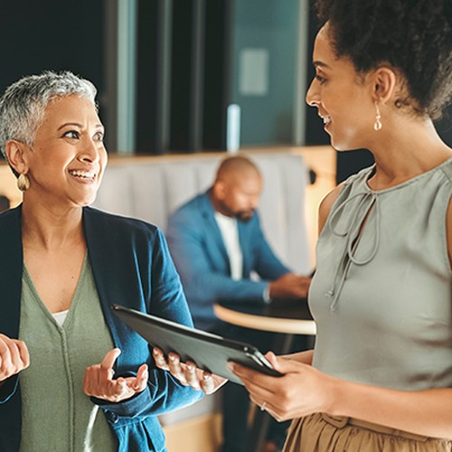 Two women converse happily, with one holding a tablet. In the background, a man focuses on a device. The setting appears modern and casual, suggesting a collaborative workspace.