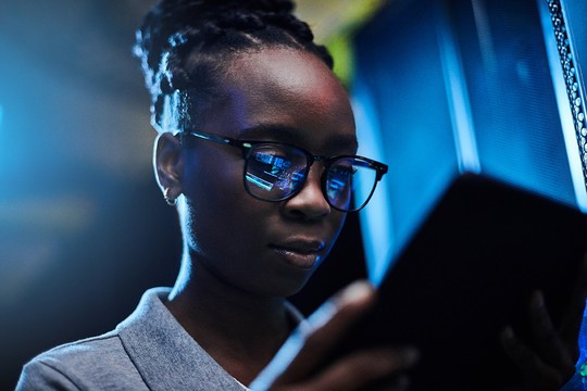A young person with glasses holds a tablet, focused on it. They are illuminated by blue light, possibly from a computer setup or digital environment in the background.