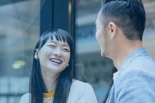 Two people are engaged in conversation, laughing and smiling. They are seated near a glass window in a bright, modern setting, with soft lighting and contemporary decor around them.