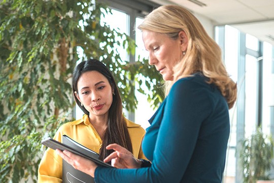 Two women are engaged in conversation, with one examining a tablet and the other listening attentively. The setting features large windows and greenery, suggesting a bright, modern office environment.