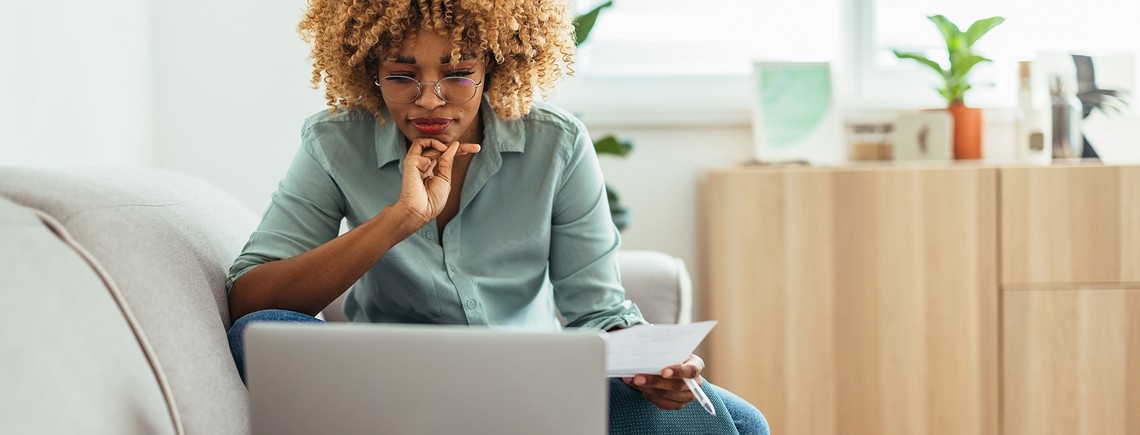 A person sits on a couch gazing at a laptop, thoughtfully holding a piece of paper. The setting is a bright room adorned with plants and light wooden furniture.