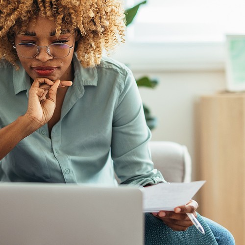 A person sits on a couch gazing at a laptop, thoughtfully holding a piece of paper. The setting is a bright room adorned with plants and light wooden furniture.