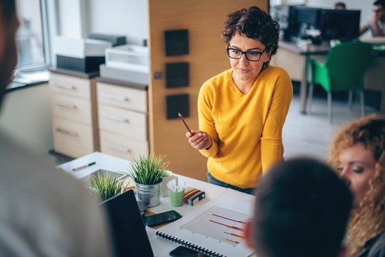 A woman in a yellow sweater engages with colleagues, holding a pencil as she discusses a chart on a table surrounded by plants, notepads, and electronics in a modern office environment.