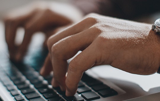 A person's hands type on a laptop keyboard, wearing a silver watch. The setting appears to be a casual workspace, with blurred background elements suggesting a comfortable environment.