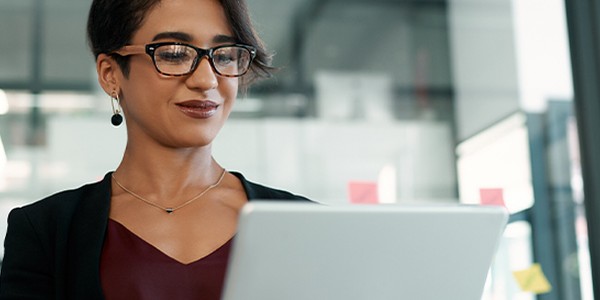 A woman with glasses is working on a laptop, smiling slightly. She is dressed in a dark blazer and a maroon top, seated in a modern office with large windows and bright natural light.