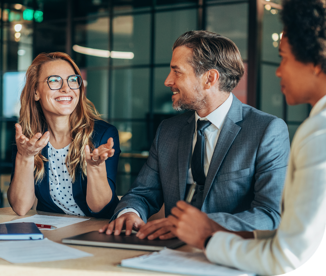 A woman gestures animatedly while speaking to two colleagues at a conference table, engaged in a professional discussion in a modern office environment with glass walls and overhead lights.