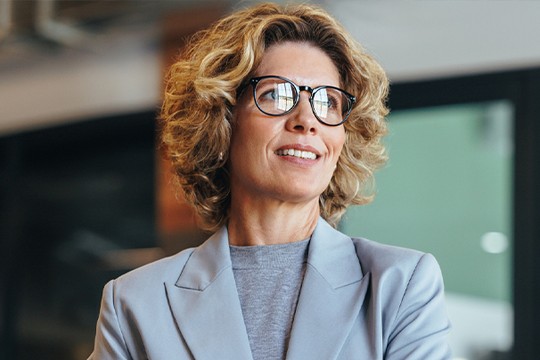 A woman with curly hair and glasses stands confidently, smiling. She wears a light blazer and looks upward, suggesting a professional or thoughtful atmosphere in a modern office space.
