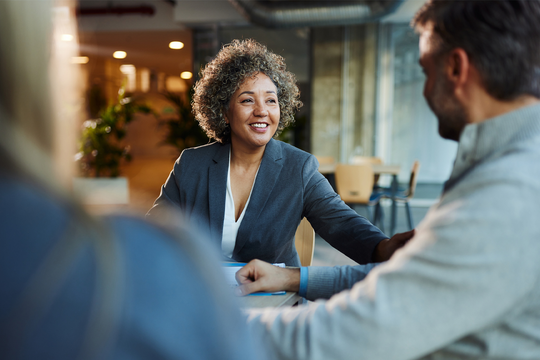A woman in a gray suit smiles and interacts with two people across a table in a modern, well-lit office space with greenery in the background.
