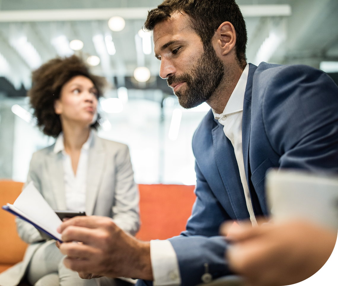 A man in a blue suit reads notes from a notepad while a woman in a gray suit looks on, both seated in an informal office setting with modern decor.