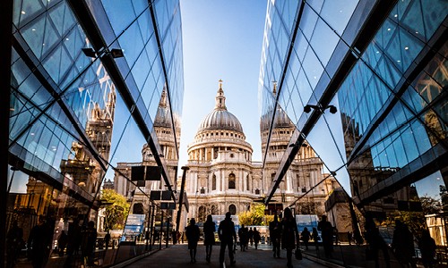 St. Paul's Cathedral stands prominently, surrounded by modern glass buildings reflecting its historic architecture. People walk towards the cathedral in a bright, clear sky, creating a stark contrast between old and new.