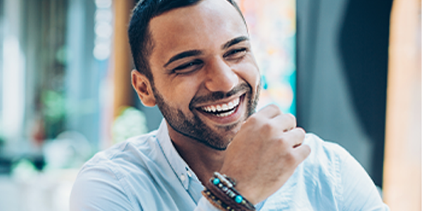 A smiling man with short hair is resting his chin on his hand, showing a joyful expression. He is in a brightly lit indoor space with greenery in the background.