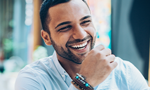 A smiling man with short hair is resting his chin on his hand, showing a joyful expression. He is in a brightly lit indoor space with greenery in the background.
