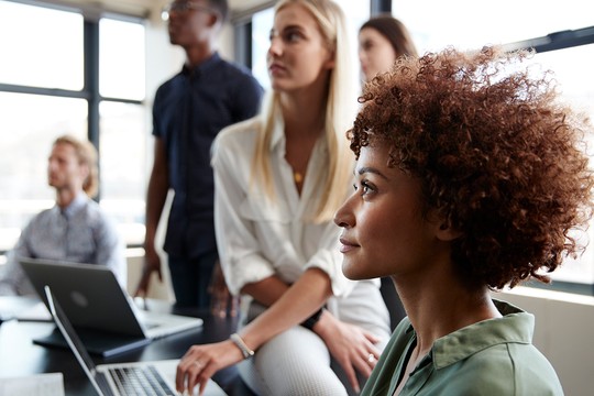A woman with curly hair is engaged in a discussion with colleagues in a modern office. Laptops are open on the table, and others are attentively listening in a bright environment.