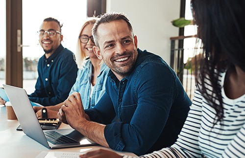 A group of four people sit around a table with a laptop open. One man smiles while engaging with others, who appear relaxed and engaged in conversation, in a bright, modern workspace.