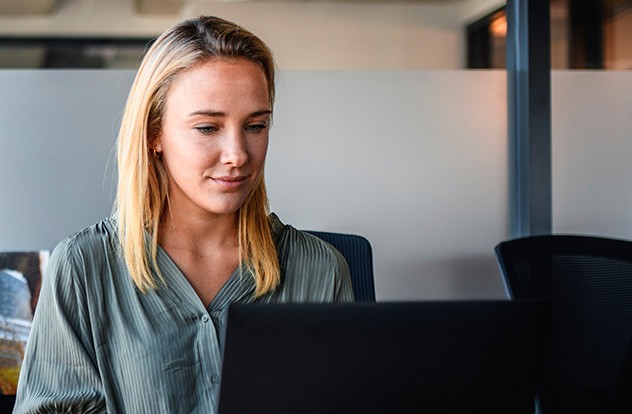 A blonde woman with straight hair sits at a desk, focused on her laptop screen. The modern office environment features glass partitions and stylish furniture, creating a professional atmosphere.