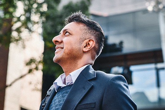 A man in a formal outfit stands with a slight smile, looking upward against a backdrop of blurred greenery and a modern building, suggesting a moment of reflection or aspiration.