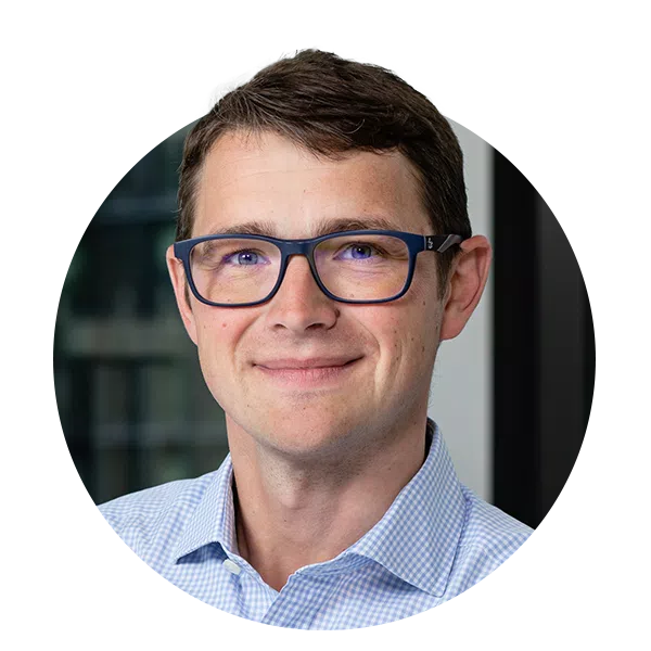 A person with short, dark hair wearing glasses and a collared shirt smiles for the camera. The background features blurred glass, suggesting an office environment.
