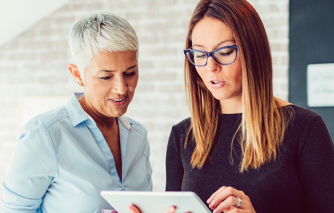 Two women engage closely with a tablet, discussing information. One wears a light blue blouse, the other in a dark sweater, in a modern, softly lit interior with brick walls.