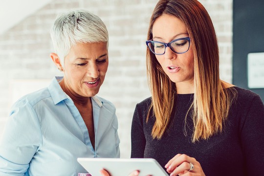 Two women engage closely with a tablet, discussing information. One wears a light blue blouse, the other in a dark sweater, in a modern, softly lit interior with brick walls.