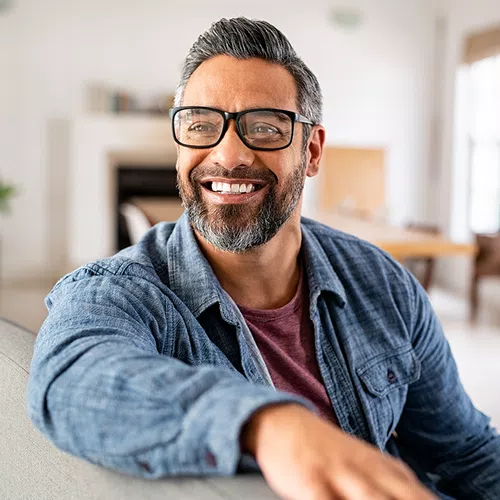 A man with glasses and a beard smiles while sitting on a couch in a bright, modern living room, with a fireplace and a table visible in the background.