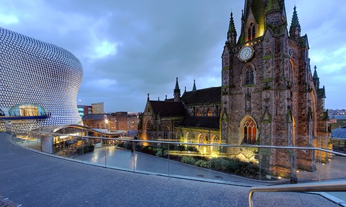 A historic stone church with a clock stands beside a modern, curved glass and metal building. The scene captures a dusk atmosphere with cloudy skies and urban surroundings.