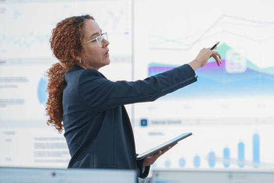 A woman in business attire points with a pen while holding a tablet, presenting data visualizations on large screens in a modern office setting focused on analytics and growth.