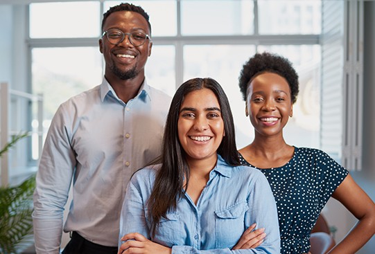 Three smiling individuals stand close together in a bright office environment with large windows. They are dressed casually, exuding a friendly and professional atmosphere.