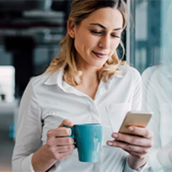 A woman holding a teal mug checks her phone while smiling. She stands by a large window in a modern office environment, with soft lighting and industrial decor.