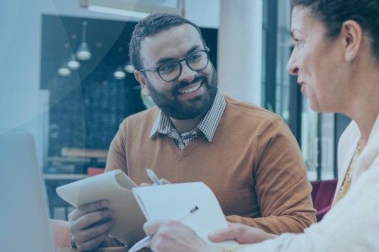 A man and woman engage in a conversation at a table. The man holds a notebook and smiles, while the woman writes notes. A modern, bright office environment is visible in the background.