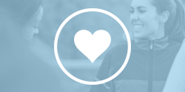A heart symbol overlays two women smiling and interacting warmly. They are outdoors, with a blurred background suggesting a casual, friendly atmosphere, possibly during a group activity or exercise.