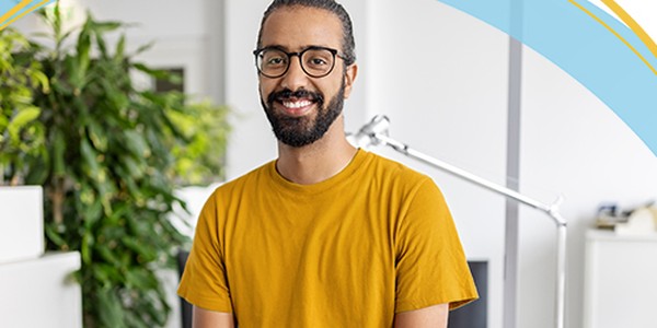 A man with a beard, wearing a yellow shirt and glasses, smiles while standing in a bright office space filled with greenery and modern furniture, suggesting a casual, professional environment.