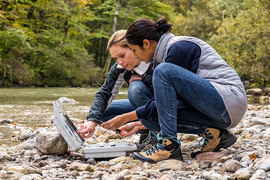 Two women crouch beside a riverbank, examining a portable device open on a flat rock. They wear casual outdoor clothing against a backdrop of trees and flowing water.