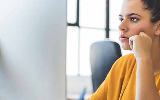 A woman in a yellow top rests her chin on her hand while focused on a computer screen. The office environment includes other desks and large windows with natural light.