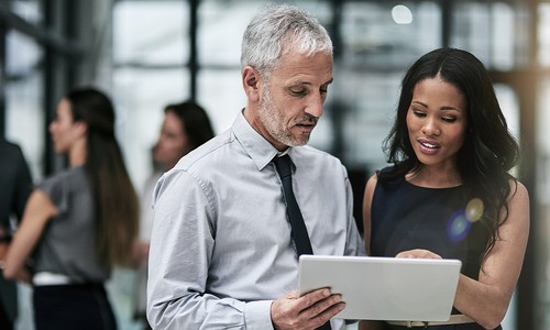 A man in a light blue shirt and tie closely examines a tablet, while a woman in a dark dress points at the screen. They are in a modern, bright office environment with colleagues in the background.
