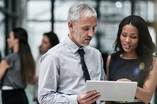 A man in a light blue shirt and tie closely examines a tablet, while a woman in a dark dress points at the screen. They are in a modern, bright office environment with colleagues in the background.