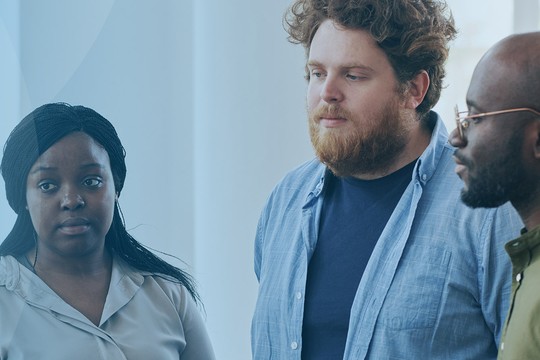 Four individuals are engaged in conversation. They appear to be discussing something important in a bright, modern environment with large windows and soft lighting.