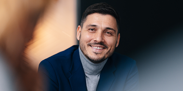 A man in a dark suit and turtleneck smiles while engaged in conversation. The setting appears modern and professional, suggesting a meeting or discussion.