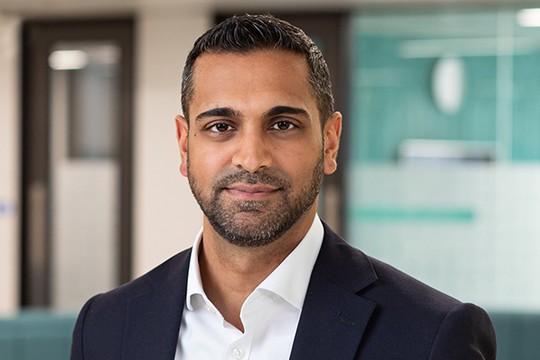 A man in a dark suit and white shirt stands centered, looking directly at the camera. He is in a modern office environment, with blurred glass and abstract green elements in the background.