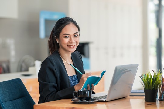 A woman in a blazer sits at a desk, smiling while writing in a blue notebook. A laptop, coffee cup, and potted plant are beside her in a bright workspace.